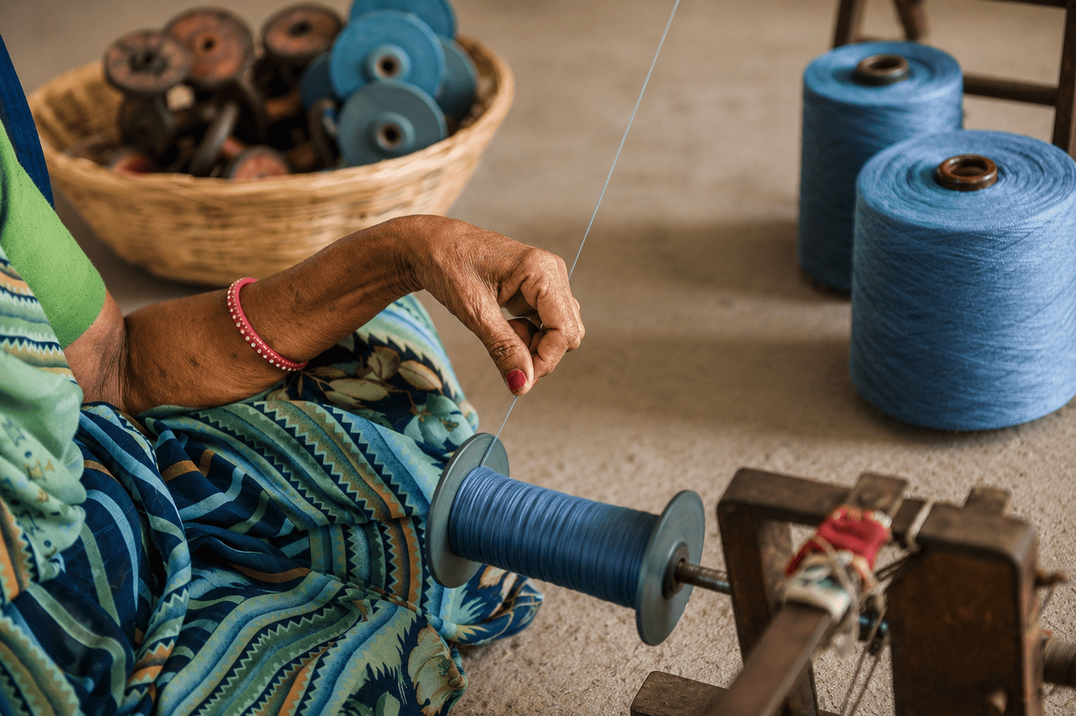 An Indian artisan woman sitting on the floor and handspinning indigo-dyed thread onto a bobbin using a traditional mechanical winder surrounded by spools of blue yarn