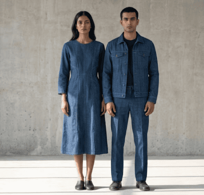 An Indian male model wearing a Khadi denim jacket and trousers standing alongside a female model wearing a Khadi denim midi dress, centered against a minimalist gray concrete wall